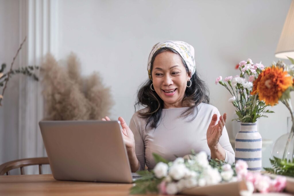 The Complete Guide to The Knot Vendor Pricing 3 A woman wearing a headscarf smiles and gestures happily while video chatting on a laptop at a table decorated with flowers in vases, possibly discussing venue managers or the knot vendor pricing for her upcoming celebration.