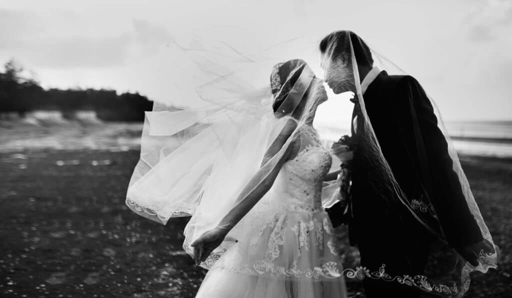 Wedding Venue Instagram Captions: 65 Ideas to Attract More Couples 3 A bride and groom stand close together on a beach, their faces nearly touching and covered by the bride’s flowing veil. The scene is romantic and windy, making it perfect for dreamy wedding venue Instagram captions with the ocean in the background.