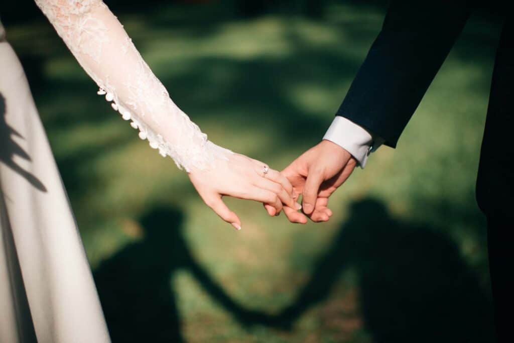 Wedding Venue Instagram Captions: 65 Ideas to Attract More Couples 2 A close-up of a bride and groom holding hands outdoors, their fingers gently intertwined, with their shadows on the grass forming a heart—perfect inspiration for funny wedding captions at your dream wedding venue.