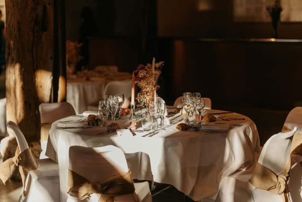 Wedding Venue Instagram Captions: 65 Ideas to Attract More Couples 1 A round table set for a formal event with white tablecloth, elegant glassware, plates, cutlery, rolls, and dried floral centerpieces. Perfect inspiration for wedding venue Instagram captions, with sunlight highlighting the charming burlap bows.