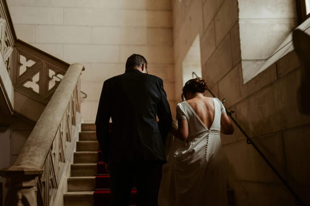 Wedding Venue Instagram Captions: 65 Ideas to Attract More Couples 4 A couple, dressed in formal attire with the woman in a white gown, walk up a stone staircase in an elegant wedding venue, softly lit. Their backs are to the camera—perfect for stunning wedding venue Instagram captions.