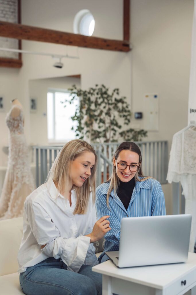 The Complete Guide to The Knot Vendor Pricing 2 Two women sit together, smiling as they look at a laptop in a bright, modern room with dresses in the background—discussing venue ideas and exploring venue managers online.