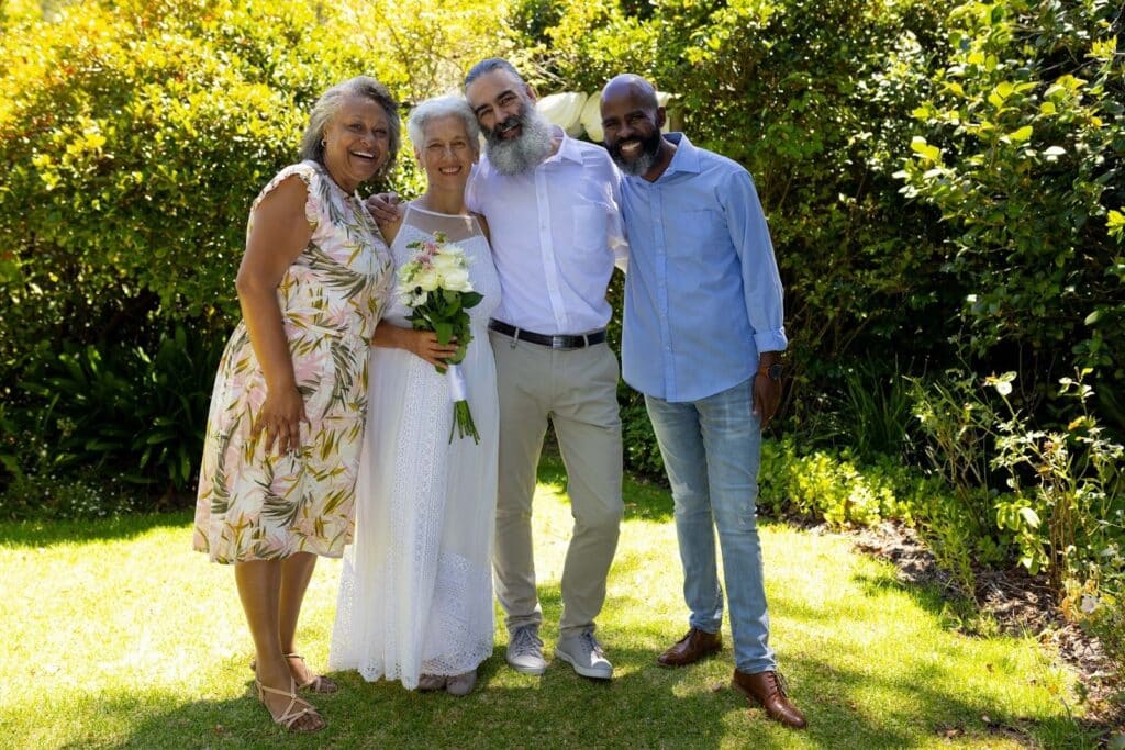 How to Attract Wedding Clients: 7 Steps to Fill Your Calendar Faster 1 Four adults stand smiling outdoors on a sunny day, with one woman in a white dress holding a bouquet of flowers—an ideal moment for any wedding photography business seeking to attract wedding clients, set against lush greenery.
