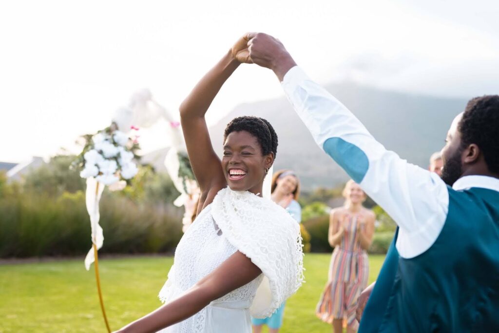 How to Attract Wedding Clients: 7 Steps to Fill Your Calendar Faster 2 A joyful bride smiles as she dances with her partner outdoors—a perfect moment for any wedding photography business. She wears a white dress and shawl; he wears a blue vest. Blurred guests and floral arrangements fill the sunny background.