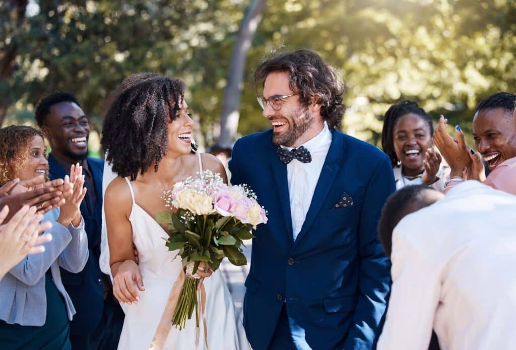 How to Attract Wedding Clients: 7 Steps to Fill Your Calendar Faster 3 A joyful bride and groom walk outdoors, smiling and holding a bouquet, as friends clap and celebrate their wedding—an ideal scene for those learning how to attract wedding clients or grow a wedding photography business. Sunlit trees complete the backdrop.