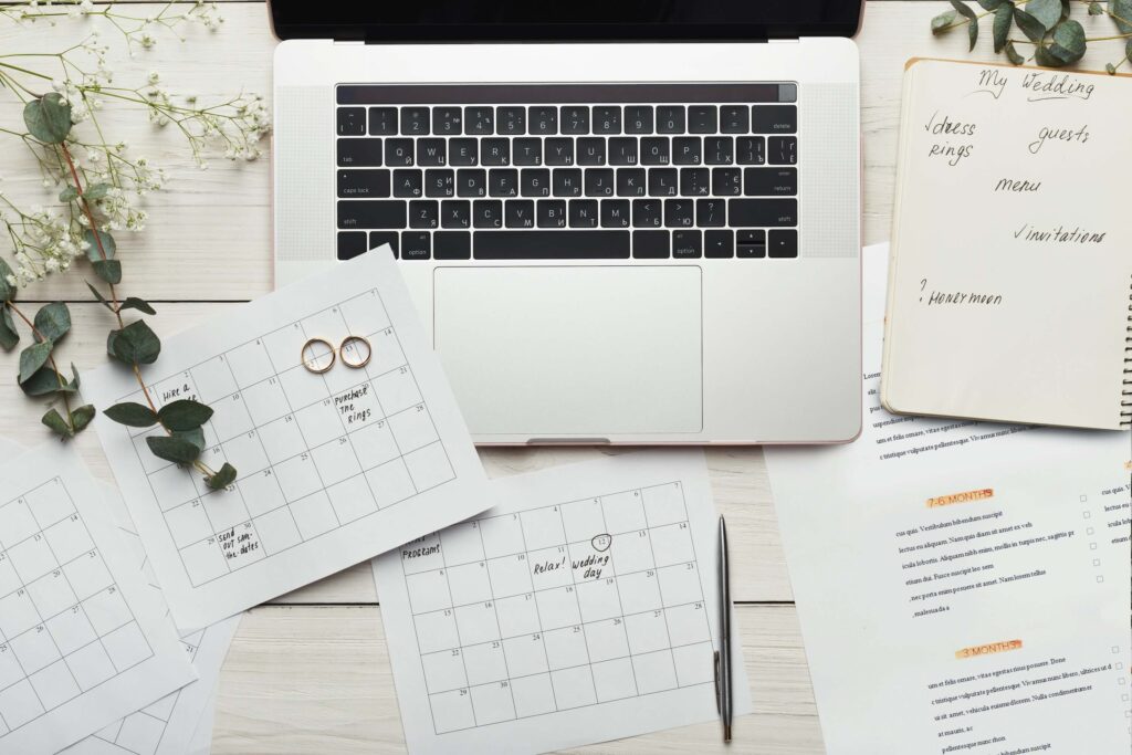 The Complete Guide to The Knot Vendor Pricing 1 A laptop on a wooden desk is surrounded by calendar pages, a pen, two wedding rings, a clipboard with wedding notes, a printed document on The Knot vendor pricing, and sprigs of greenery—perfect for planning your venue.