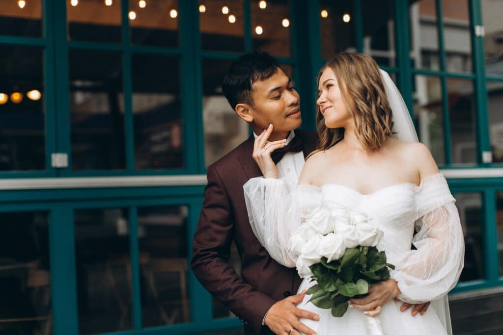 Wedding Venue Industry Analysis in 2026 and Beyond 4 A bride in a white dress and veil holding a bouquet of white roses stands close to a groom in a dark suit and bow tie. They gaze lovingly at each other, capturing the magic of a destination wedding in front of a blue windowed building.