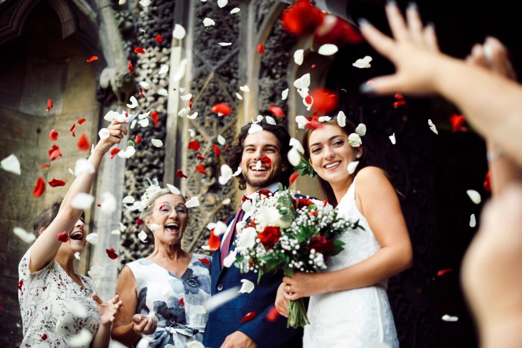 Owning a Wedding Venue: What It Takes to Start, Open, and Run a Profitable One 8 A newlywed couple smiles joyfully outside, holding a bouquet, as guests throw red and white flower petals into the air—capturing the magic of owning a wedding venue where every moment feels happy and festive.