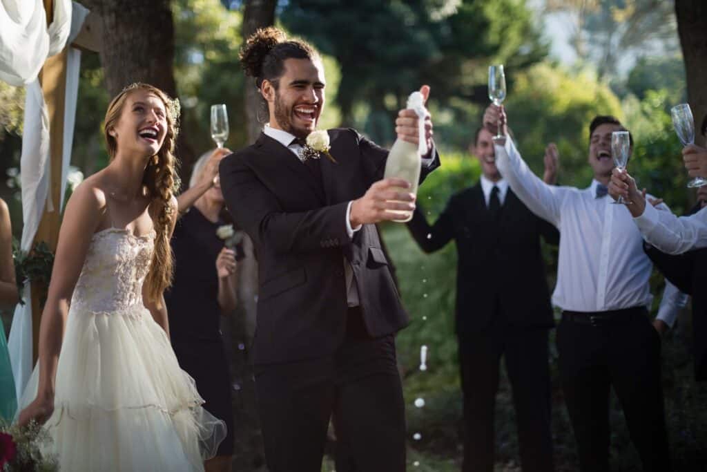 Owning a Wedding Venue: What It Takes to Start, Open, and Run a Profitable One 3 A bride and groom celebrate outdoors with friends, popping a bottle of champagne. The bride smiles in a white dress, while the groom, in a black suit, opens the bottle as guests cheer and hold up champagne glasses.