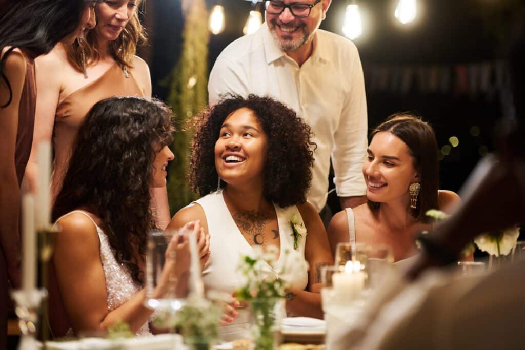 Wedding Venue Industry Analysis in 2026 and Beyond 5 A group of people dressed in formal attire smile and talk around a dinner table at a destination wedding, decorated with candles and greenery, under warm string lights at an outdoor evening event.