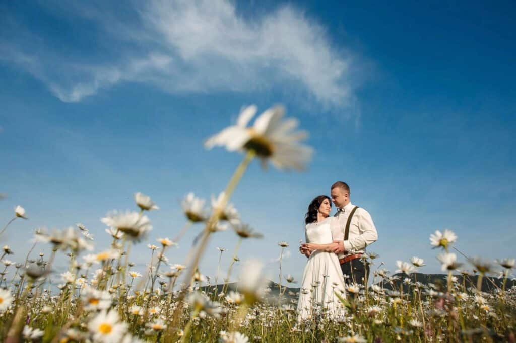 Owning a Wedding Venue: What It Takes to Start, Open, and Run a Profitable One 1 A couple stands embracing in a field of daisies under a bright blue sky with wispy clouds. The woman wears a white dress and the man is in a white shirt with suspenders. Some daisies are blurred in the foreground.