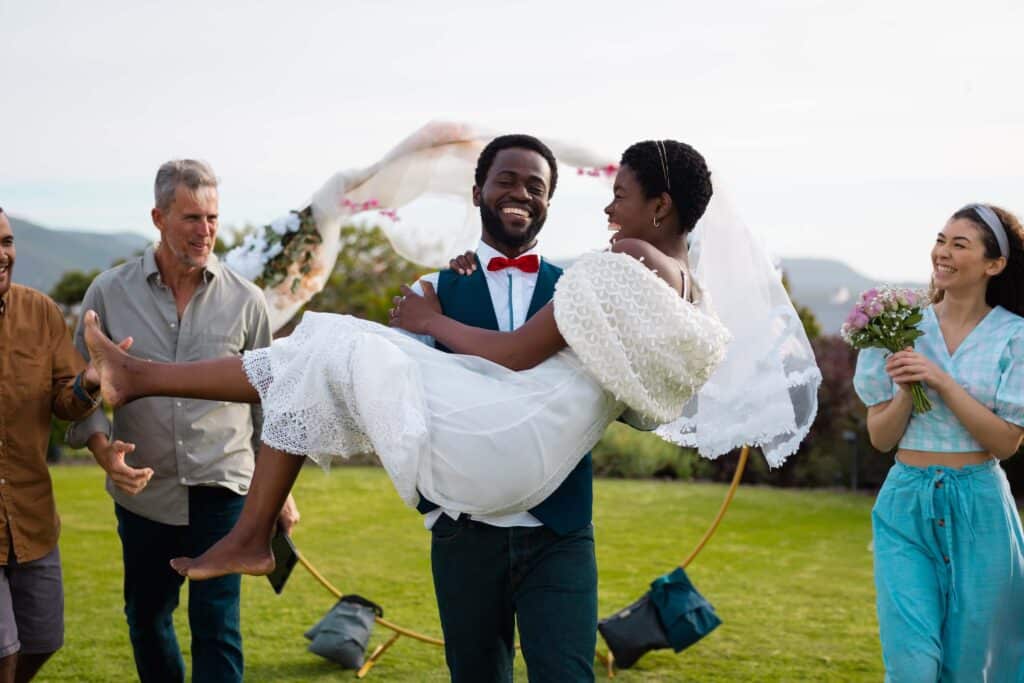 Owning a Wedding Venue: What It Takes to Start, Open, and Run a Profitable One 2 A groom in a suit and red bow tie joyfully carries his smiling bride in a white dress and veil outdoors, surrounded by happy friends celebrating at a wedding.
