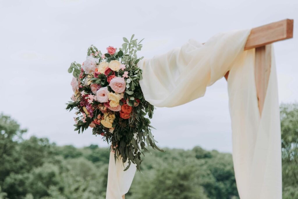 Wedding Venue Business Model: How to Build a Thriving, Sustainable Venue Business 1 A floral arrangement with pink, peach, and white flowers and greenery adorns a wooden arch draped in sheer ivory fabric—an inspiring setting for your wedding venue business plan, nestled outdoors with trees in the background.