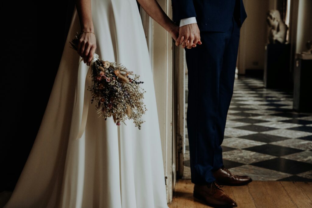 Wedding Venue Industry Analysis in 2026 and Beyond 1 A bride in a white dress holding a bouquet stands next to a groom in a dark suit; they are holding hands, partially visible, on the checkered floor of an elegant destination wedding.