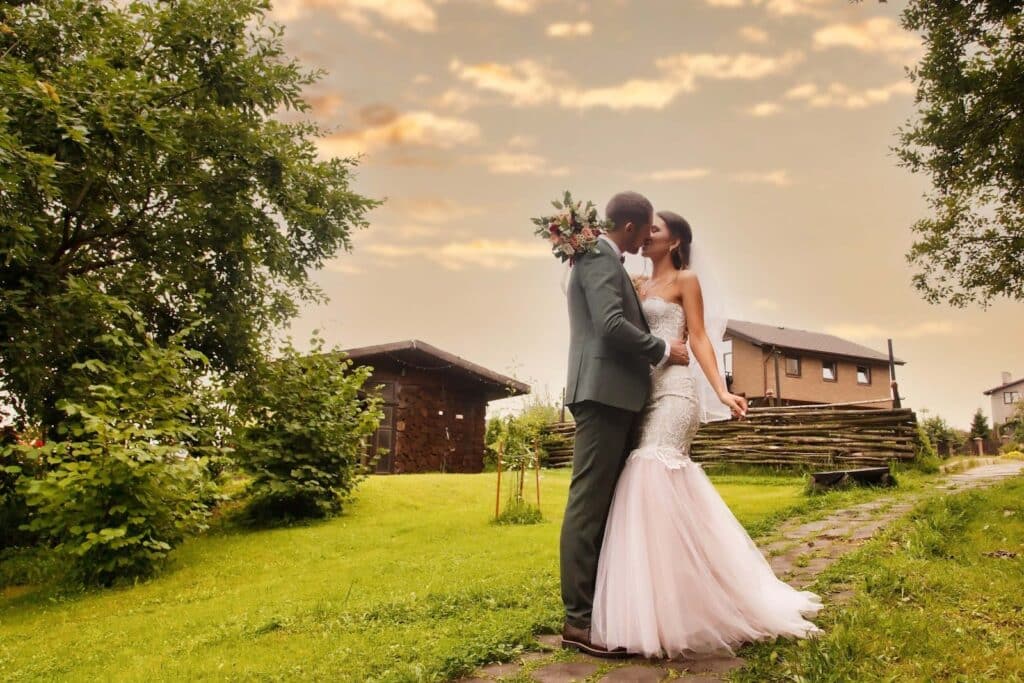 Wedding Venue Business Model: How to Build a Thriving, Sustainable Venue Business 4 A bride and groom stand close together on a stone path in a lush, green garden at sunset—an idyllic wedding venue with rustic wooden houses and a glowing sky, perfect inspiration for any wedding venue business plan.