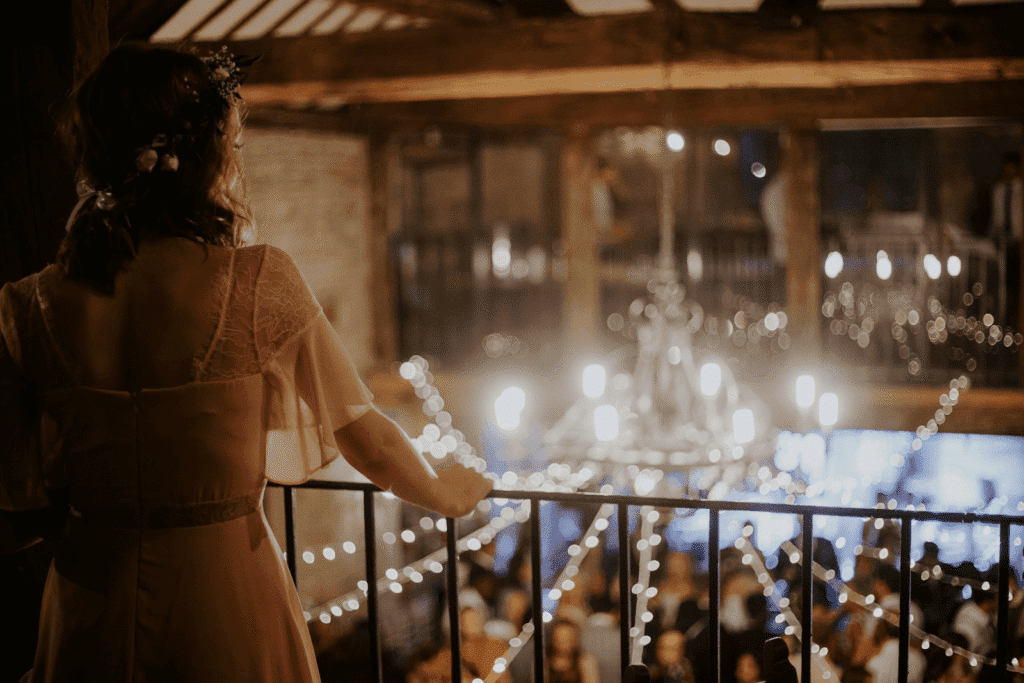 Wedding Venue Business Model: How to Build a Thriving, Sustainable Venue Business 3 A woman in a light dress stands on a balcony, overlooking a warmly lit, crowded wedding venue decorated with string lights and a chandelier, creating a festive, elegant atmosphere.
