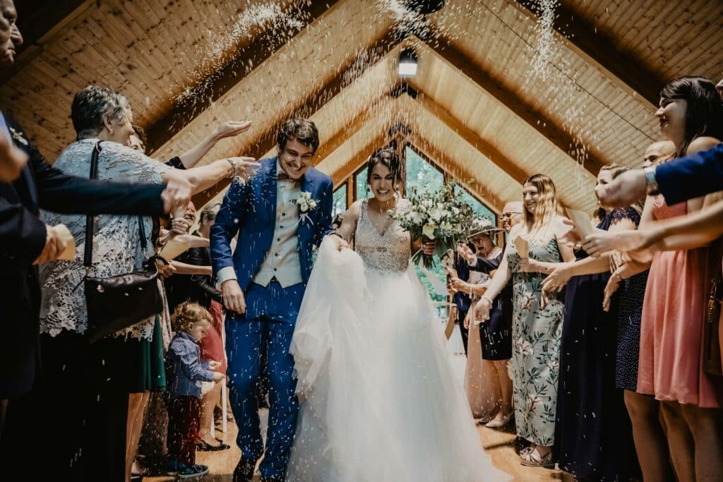 Wedding Venue Industry Analysis in 2026 and Beyond 2 A joyful bride and groom walk down the aisle in a wooden hall, as guests from the wedding venue industry shower them with rice or confetti, celebrating their wedding with smiles and applause all around.