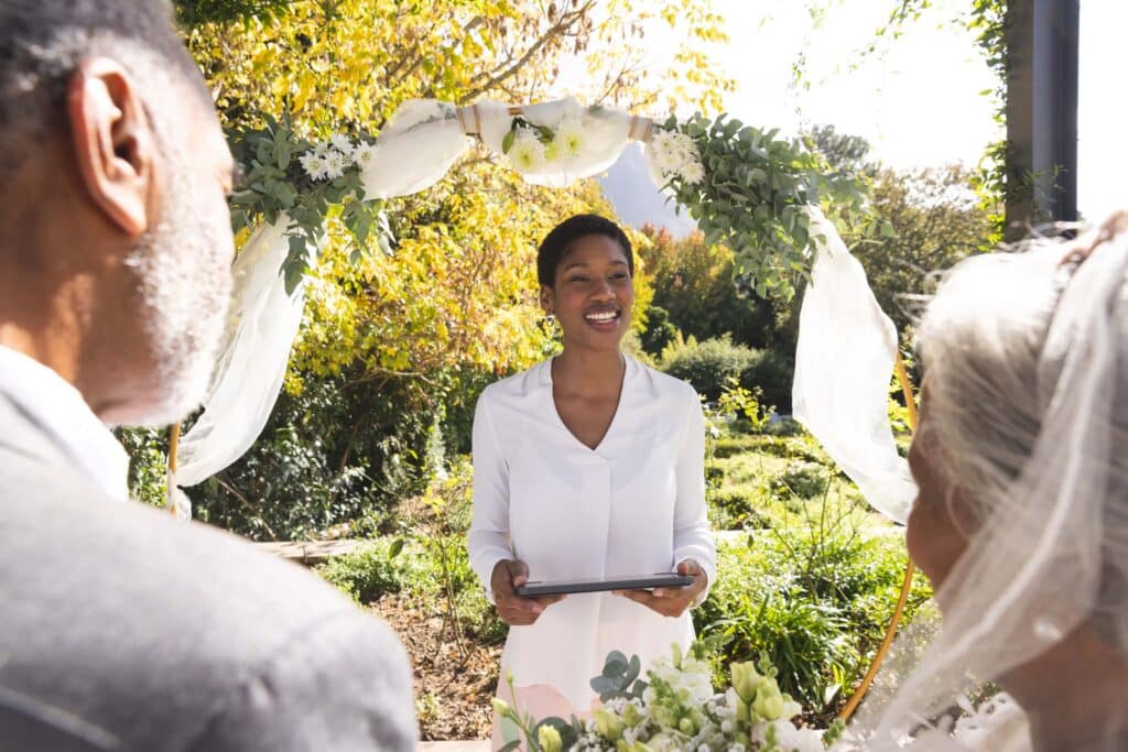 Is the Wedding Industry Dying in 2026? A Clear Look at What’s Changing 1 A woman officiates an outdoor wedding ceremony, smiling at a couple before her. The scene showcases a floral arch and greenery in the background, with sunlight filtering through—highlighting the dedication of wedding professionals.
