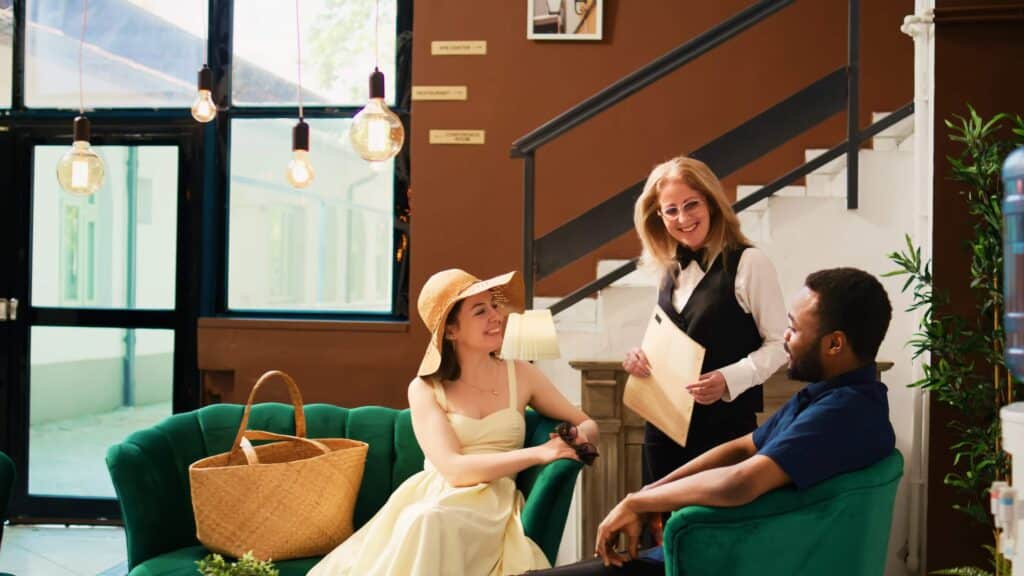 The Top Wedding Industry Trends for 2026 + 6 Tips for Venue Owners 2 A smiling hotel receptionist greets a seated couple in a cozy lobby, perhaps discussing wedding venue options. The woman wears a summer dress and hat, the man sits beside her, with a straw bag on the green couch and a staircase in the background.