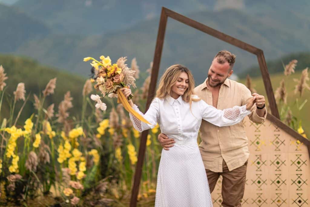 The Top Wedding Industry Trends for 2026 + 6 Tips for Venue Owners 6 A smiling couple poses outdoors at a stunning wedding venue; the woman in a white dress holds yellow flowers, embraced by her partner. Behind them are yellow blooms and mountains, capturing timeless romance in the heart of the wedding industry.