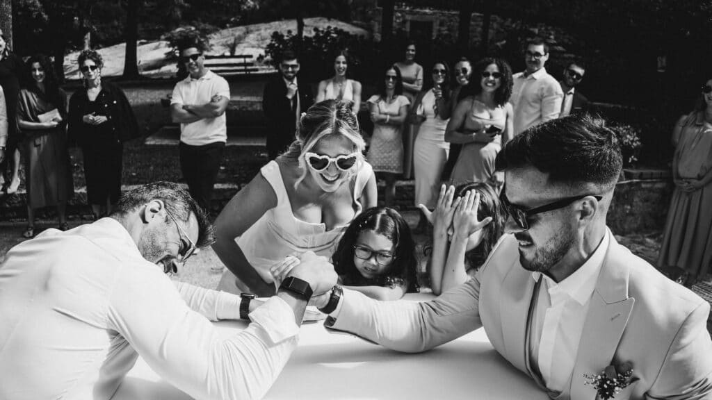 The Top Wedding Industry Trends for 2026 + 6 Tips for Venue Owners 1 Two men arm wrestling at an outdoor wedding venue, surrounded by a cheering group. A woman in heart-shaped sunglasses and a girl watch closely, with others smiling nearby—a lively, festive scene capturing fun wedding industry trends.
