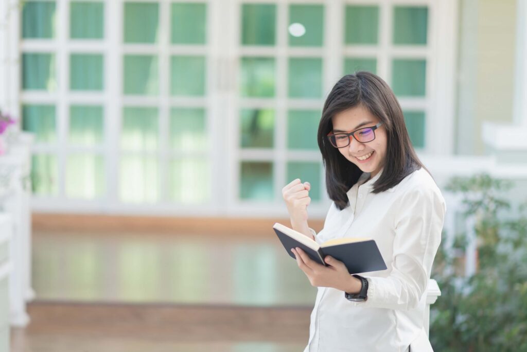 Is the Wedding Industry Dying in 2026? A Clear Look at What’s Changing 4 A young woman wearing glasses and a white shirt smiles and raises her fist in excitement while reading a book about wedding professionals, standing outdoors in front of a building with large windows.