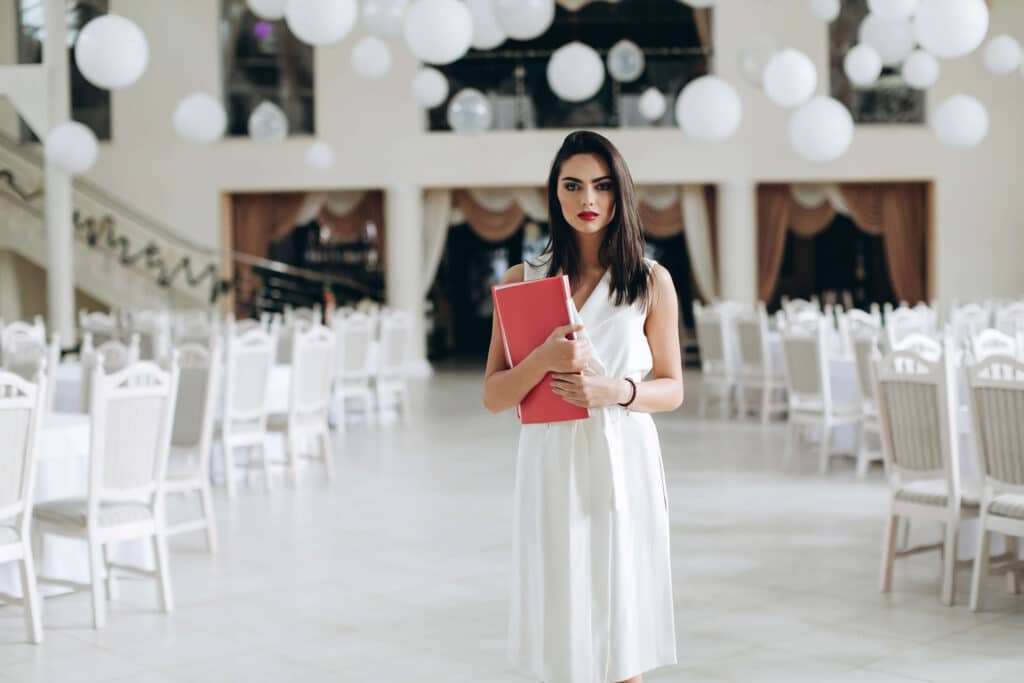 Is the Wedding Industry Dying in 2026? A Clear Look at What’s Changing 12 A woman in a white dress stands in an elegant, white-decorated event hall, holding a red folder. With the bright room full of white chairs and round decorations, one may wonder: is the wedding industry dying?