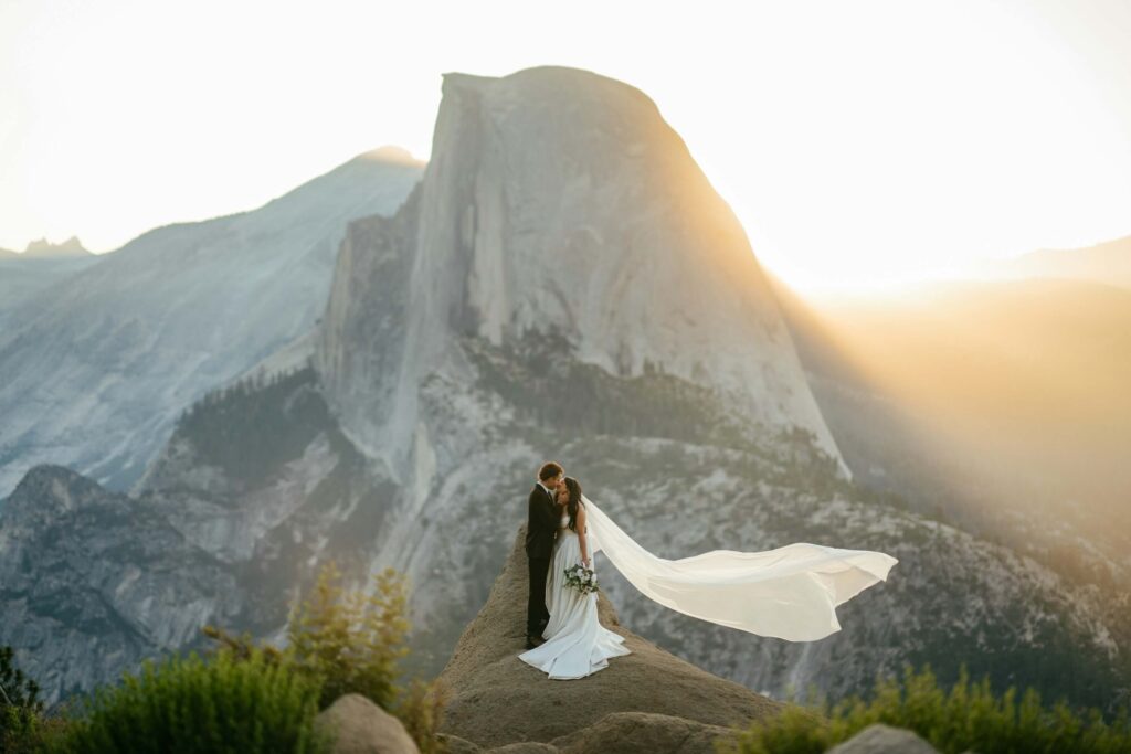 The Top Wedding Industry Trends for 2026 + 6 Tips for Venue Owners 5 A bride and groom kiss on a rocky ledge at sunset—an unforgettable moment that captures wedding industry trends. Her veil flows in the wind, framed by majestic mountains and Half Dome glowing in golden light—nature’s ultimate wedding venue.