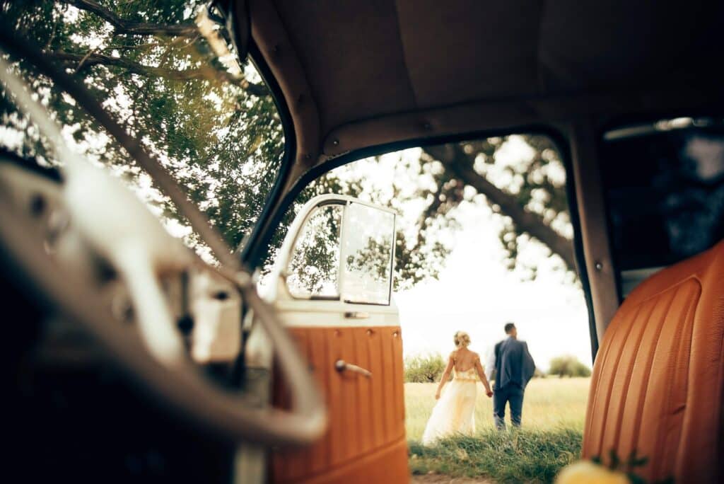 The Top Wedding Industry Trends for 2026 + 6 Tips for Venue Owners 3 A bride and groom walk hand-in-hand through a grassy field, seen through the open door of a vintage car. This romantic scene captures one of the latest wedding industry trends, highlighting unique wedding venue inspiration.