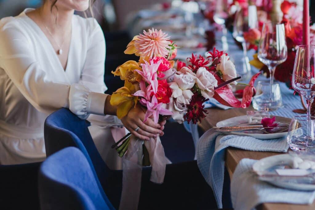 Wedding Venue Business Plan (Free Template) + 7 Areas Most Owners Overlook 1 A person in a white blouse holds a colorful bouquet of flowers while sitting at a table set with glasses, plates, and napkins, decorated with more flowers.