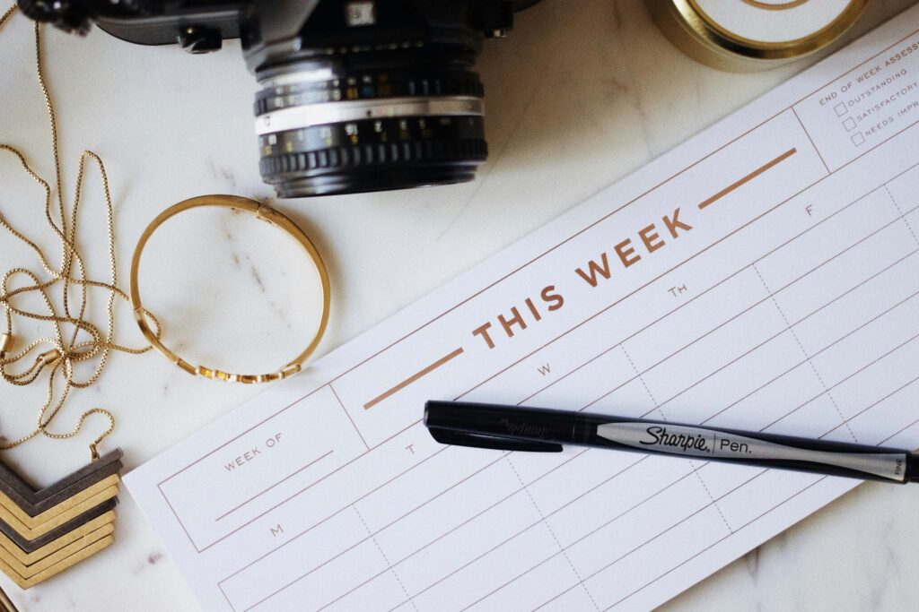 Wedding Business Branding for Venue Owners: 4 Tips That Actually Hold Up 2 A weekly planner sheet titled "THIS WEEK" lies on a white surface with a Sharpie pen on top. Nearby are a camera, a gold bracelet, a necklace, and a roll of washi tape.