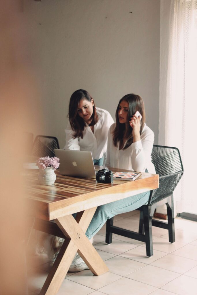 Wedding Venue Business Plan (Free Template) + 7 Areas Most Owners Overlook 2 Two women in white shirts work together at a wooden table with a laptop, camera, and flowers. One is sitting and talking on the phone, while the other stands beside her, leaning over to look at the screen.