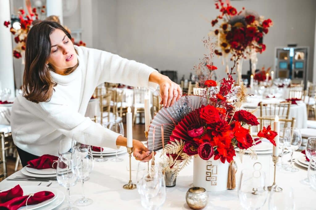 Wedding Venue Business Plan (Free Template) + 7 Areas Most Owners Overlook 4 A woman in a white sweater arranges a centerpiece of vibrant red flowers and decorative fans on a formal dining table set with glassware, candles, and gold chairs in an elegant venue.