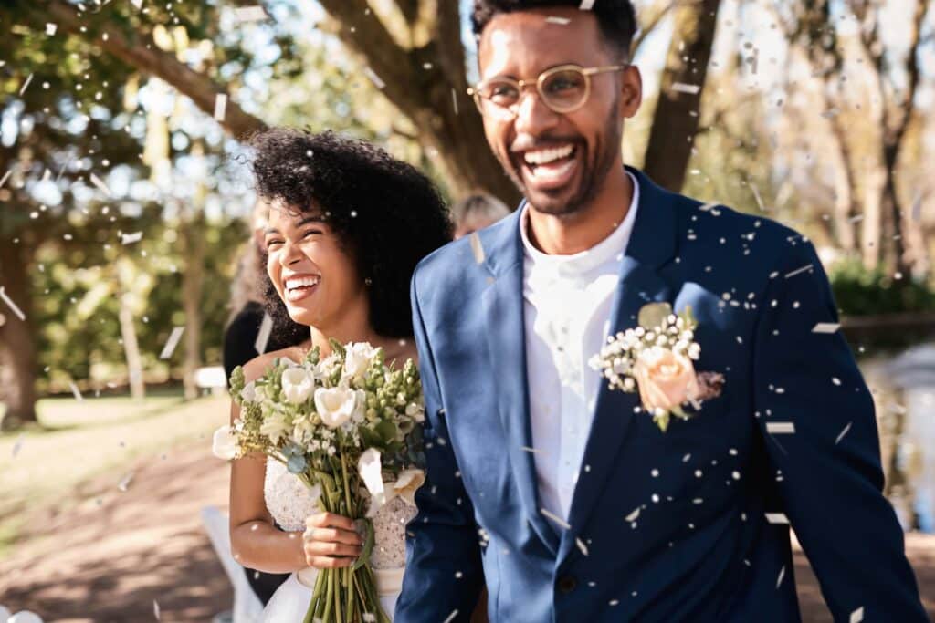 Wedding Business Branding for Venue Owners: 4 Tips That Actually Hold Up 1 A joyful bride and groom smile as they walk outdoors in sunlight, surrounded by falling confetti. The bride holds a bouquet of white flowers, and the groom wears a blue suit with a boutonniere.