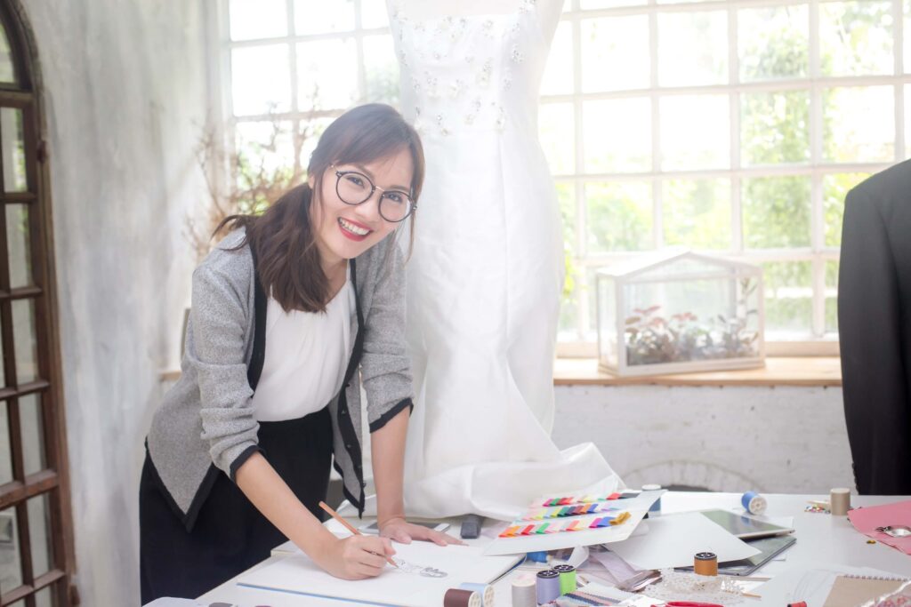 Wedding Venue Business Plan (Free Template) + 7 Areas Most Owners Overlook 11 A smiling woman wearing glasses stands at a desk, sketching designs for a wedding venue business plan. Behind her is a white dress on a mannequin, and art supplies, threads, and fabrics cover the table. Sunlight streams through large windows.