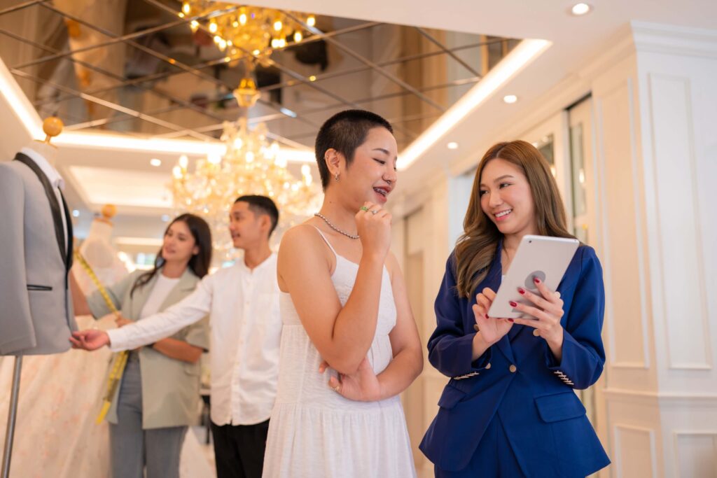 How to Run a Wedding Venue: The 5 Pillars That Keep It Booked 4 Two women stand in the foreground, one in a white dress and one in a blue blazer holding a tablet—perfectly illustrating how to run a wedding venue. In the background, others examine a suit on a mannequin in an elegant room with chandeliers.
