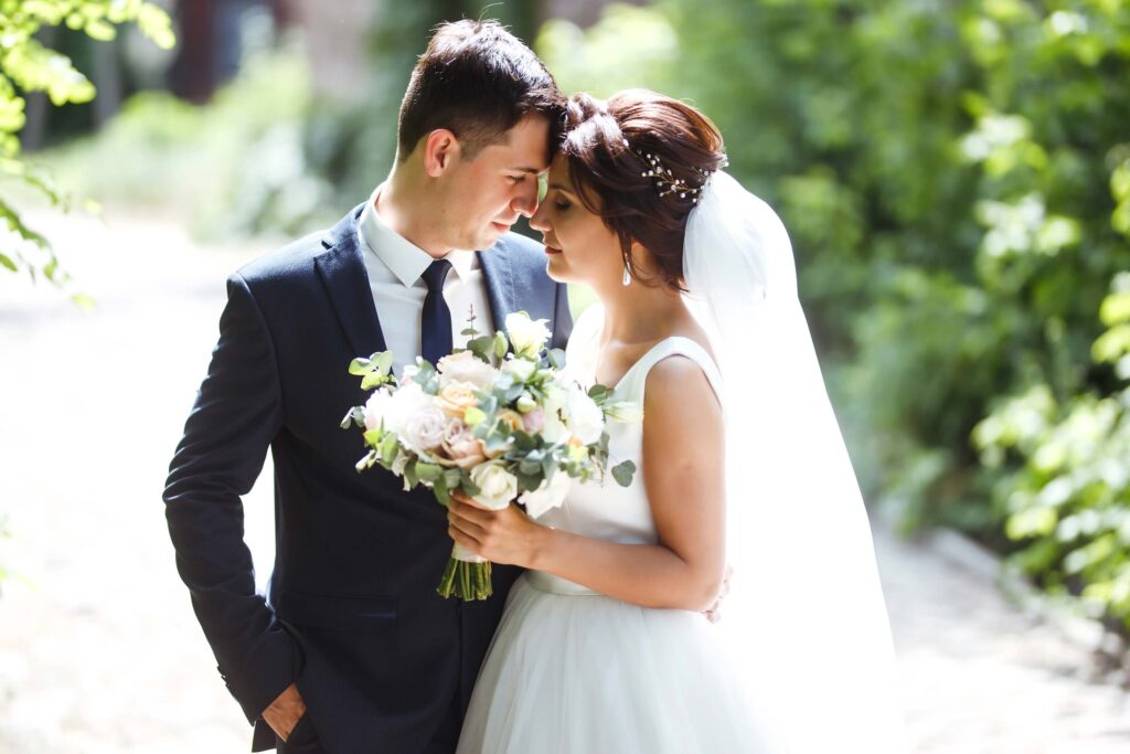 Zola vs The Knot: Which One is Right for Your Venue? 1 A bride and groom stand outdoors, embracing and touching foreheads. The bride holds a bouquet and wears a white dress with veil, while the groom wears a dark suit—capturing the magic that makes the wedding industry shine. Green foliage blurs in the background.