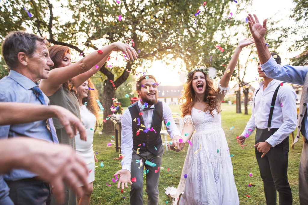 How to Run a Wedding Venue: The 5 Pillars That Keep It Booked 1 A joyful bride and groom celebrate outdoors with friends at a beautiful wedding venue, all smiling and tossing colorful confetti in the sunlit air, surrounded by trees.