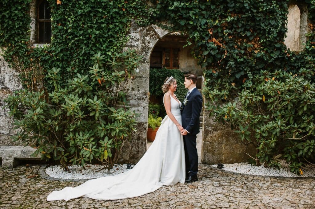 How to Run a Wedding Venue: The 5 Pillars That Keep It Booked 2 A bride in a long white gown and a groom in a dark suit stand holding hands beneath a stone archway covered in green ivy, showcasing the charm of a successful wedding venue outside a rustic building.