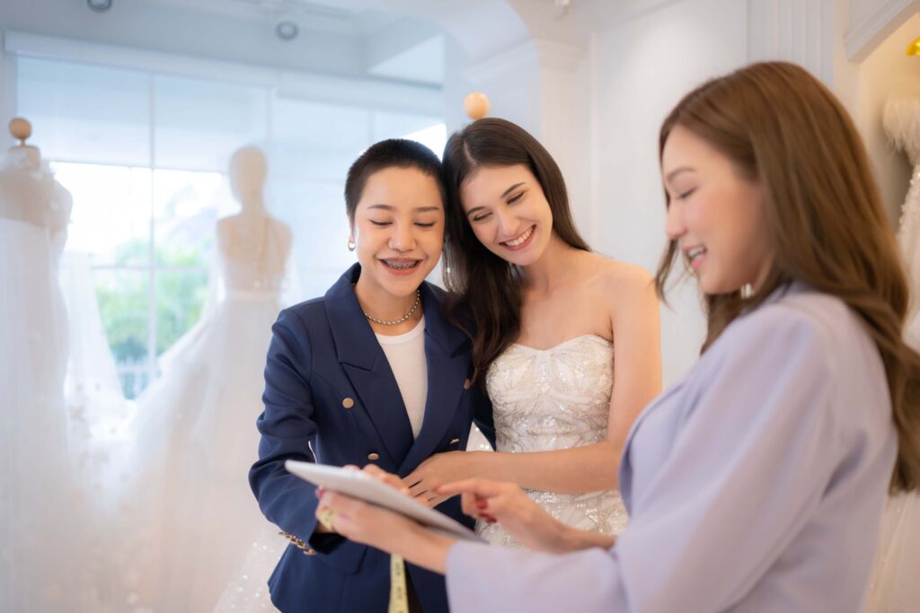 Zola vs The Knot: Which One is Right for Your Venue? 2 Three women smile together in a bridal shop, discussing free wedding ideas. One wears a wedding dress, another in a blue suit holds a notepad and pen, while the third in lavender points at the notepad. White wedding dresses fill the background.