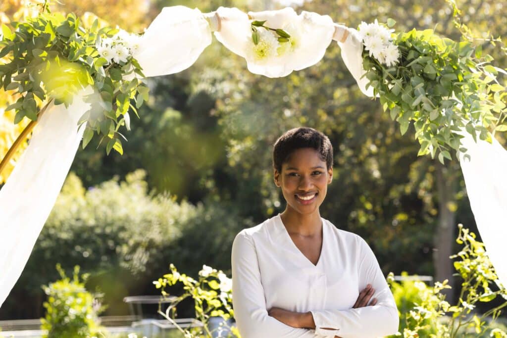 How to Run a Wedding Venue: The 5 Pillars That Keep It Booked 9 A woman in a white blouse stands smiling with arms crossed under a decorated arch, outdoors in a sunlit garden—the perfect inspiration for how to run a wedding venue with elegance and charm.