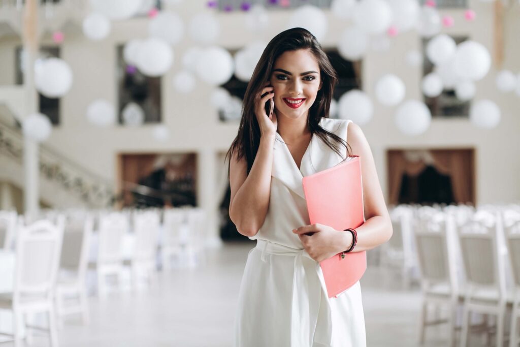 Zola vs The Knot: Which One is Right for Your Venue? 8 A woman in a white dress smiles while talking on the phone, holding a pink folder—perhaps discussing zola vs the knot. She stands in a bright room filled with white hanging ornaments and empty white chairs, embracing the excitement of the wedding industry.