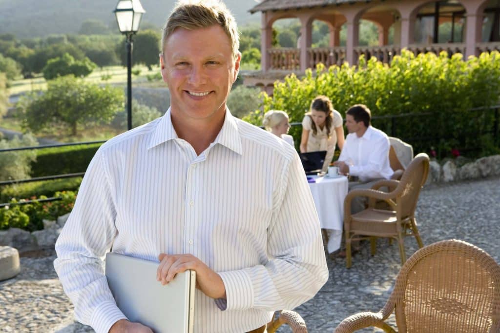 Man standing outdoors holding a laptop, representing owning a wedding venue while overseeing clients and staff at an event space