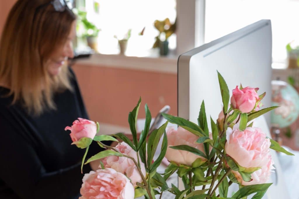 Woman working at a computer with pink floral arrangement in the foreground, representing behind-the-scenes management of wedding venue ads and marketing operations