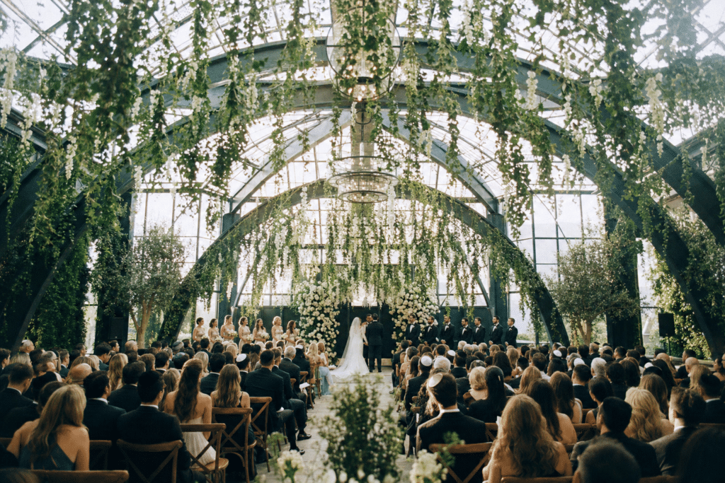 Editorial film photograph of a wedding actively in progress at a conservatory venue, couple at the altar with guests seated under hanging botanical greenery in natural daylight — the venue alive moment that the channel-mix strategy is built to fill