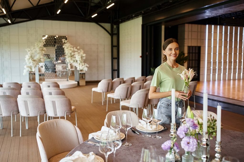 Wedding Venue Off Season Booking Strategy: Mid-Week + Shoulder-Season Plays 1 Wedding venue staff member setting tables for an off-season reception with red floral centerpieces and gold chairs.